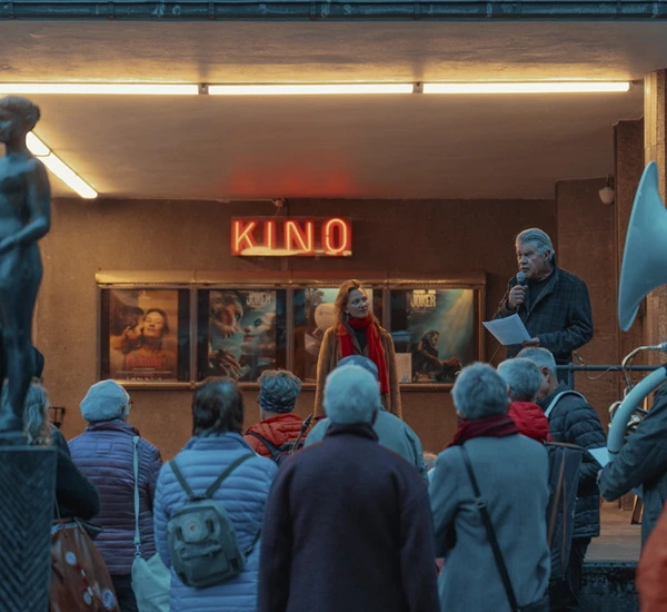 Litteraturguider Tora Døskeland og Terje Kollbotn står på trappen til Odda kino og snakker til en menge tilhørere. Neonskilt med teksten Kino i bakgrunnen.. Foto.